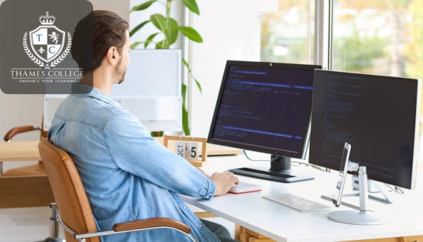 Man sitting at a desk working on dual computer monitors displaying code and data, with the Thames College logo in the corner.